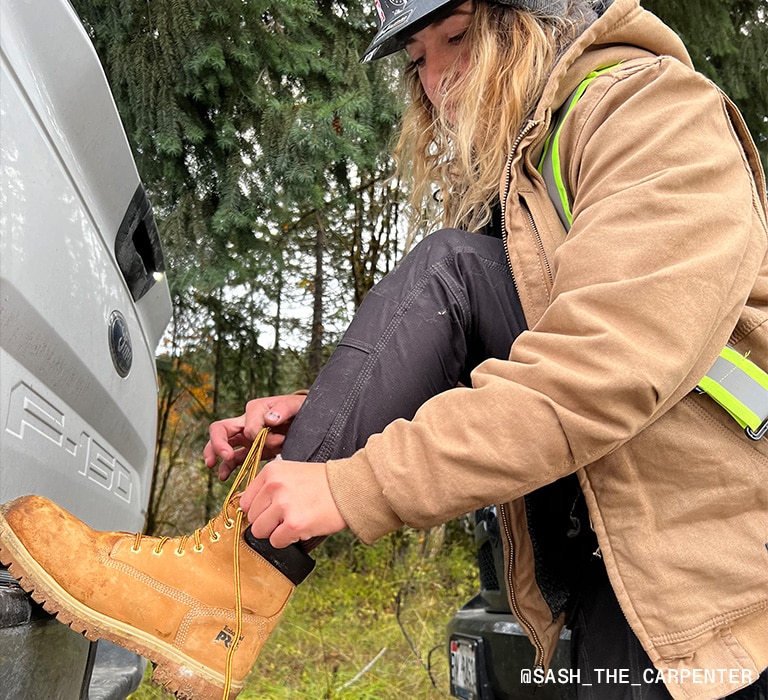 Two images of the same blond woman in a hard hat, tan work jacket, plaid shirt, black pants and wheat Timberland work boots, outside by a truck on the side of the road. In one image she's standing by the truck and in the other she's tying her boot laces while resting her foot against the tailgate.
