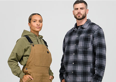Image of a woman in brown overalls and green Timberland hoodie, standing beside a man in a Timberland gray and black work flannel.