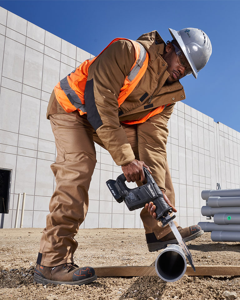 Image of a man wearing matching tan colored Timberland PRO work jacket and work pants, brown PRO work boots, with an orange safety vest, safety glasses and black hard hat. He's standing on a dirt lot outside a new concrete building that's not finished, using a saw to cut a PVC pipe. Image of a brown work jacket with black reinforced elbow patches, a brown pair of work overalls and a black pair of work cargo pants all hanging from a wooden strip of wood against a concrete background.
