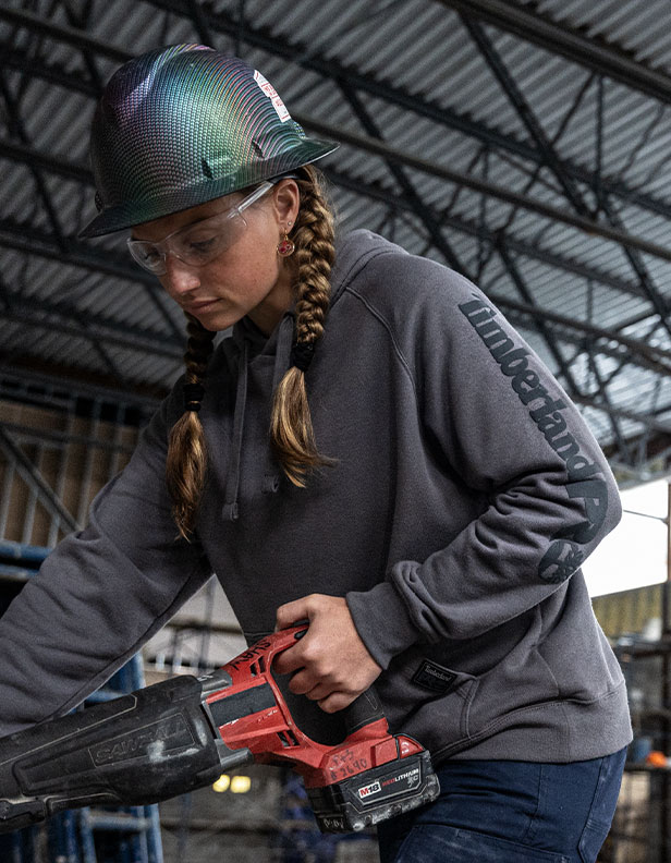 Image of a woman in brown braids standing in a warehouse on concrete, wearing a black hard hat and brown Timberland work overalls, working in a construction setting.