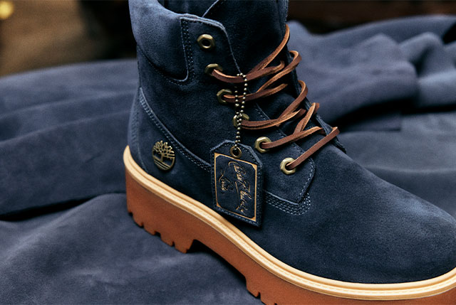 Image of extreme closeup of the side of a suede boot. The suede is a deep blue and the texture of the leather appears soft. A gold and blue hangtag saying Charles F Stead falls in elegant script. A bit of the brown outsole can be seen.