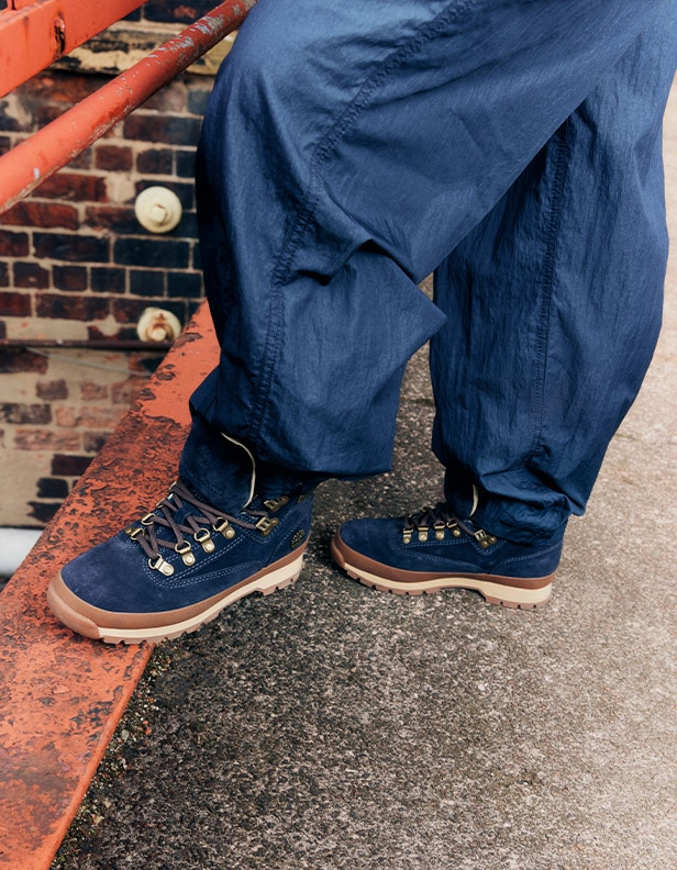 Image of a person from the knees down, standing at the top of some industrial orange metal stairs with a brick wall behind. The person is wearing blue Timberland Indigo Suede Euro Hiker boots that are fashionably tied with no visible bows.