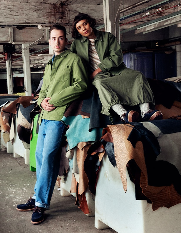 Image of a man and woman in a garment warehouse, both wearing dark blue boots from the Timberland Indigo Suede collection. They are both dressed in green jackets as he leans against a pile of dyed leathers and she sits on top of it.