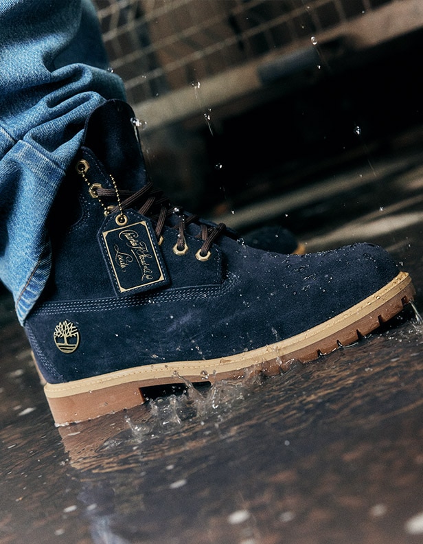 Image from just the ankle down of a person wearing jeans with the Timberland Indigo Suede 6-Inch Boot, just one foot. The boot is dark blue suede with golden accents and brown outsoles and the person is standing in a shallow puddle outside a brick building.